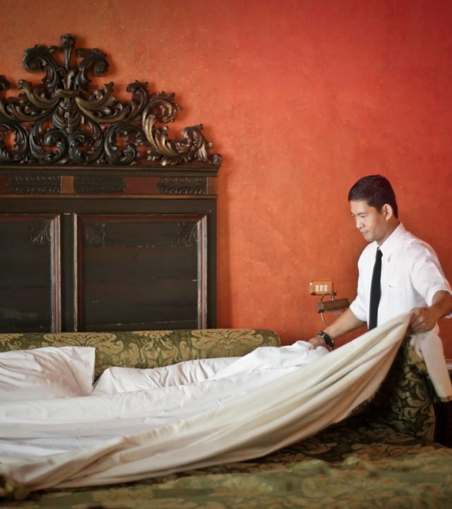Side view of young ethnic male making bed while cleaning stylish hotel room with vintage furniture and red walls