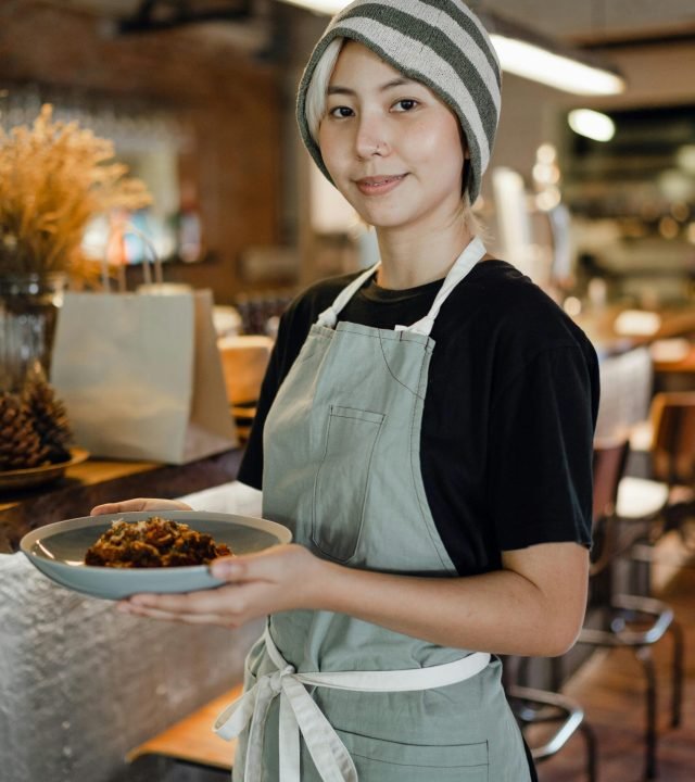 Smiling waitress standing in a cozy café holding a delicious meal, embodying hospitality and service.